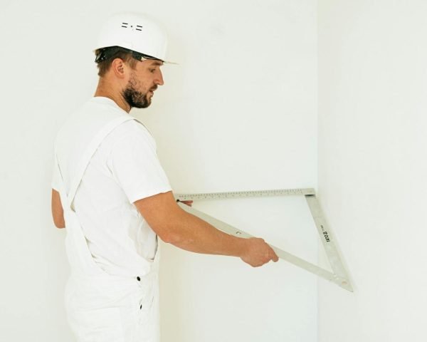 Side view of a construction worker with safety helmet measuring a wall corner indoors.